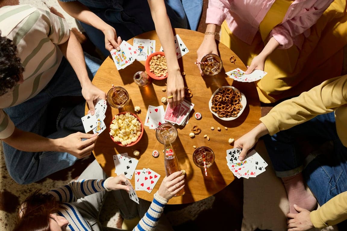 Zes vrienden rond een ronde tafel met kaarten in de hand, popcorn en drankjes ertussen